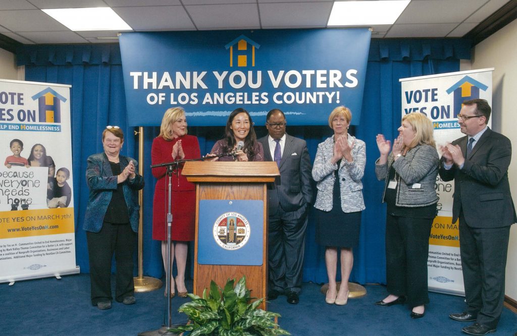 A group of LA County officials applauding at a podium, celebrating voter support for homelessness initiatives.