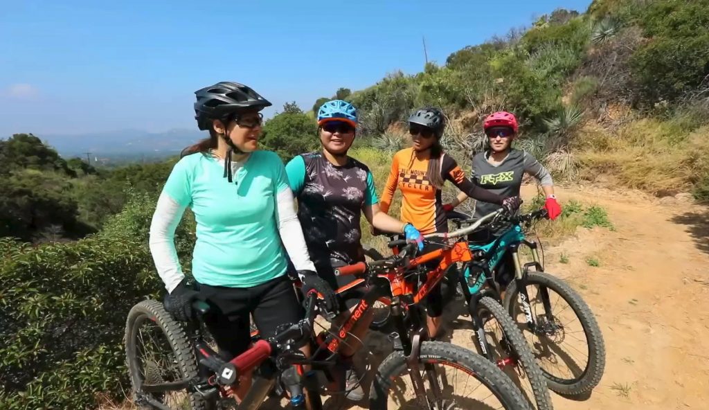 Four mountain bikers wearing helmets and cycling gear pose together on a trail surrounded by greenery.