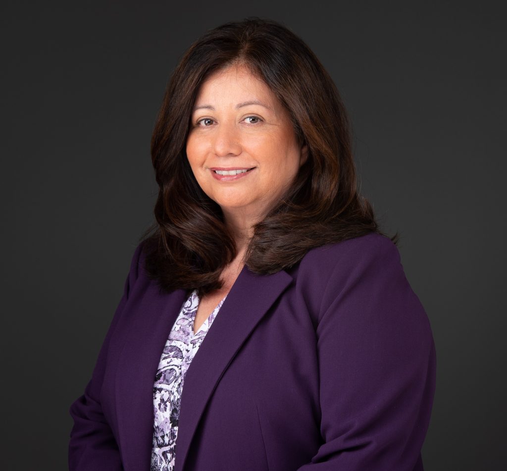 Professional portrait of a woman in a purple blazer, smiling against a dark background, conveying a sense of approachability.