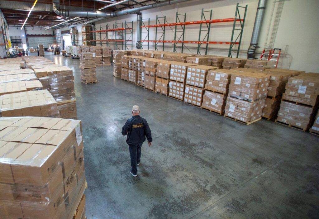 Warehouse worker walking through rows of stacked boxes, highlighting logistics for COVID-19 resource distribution.