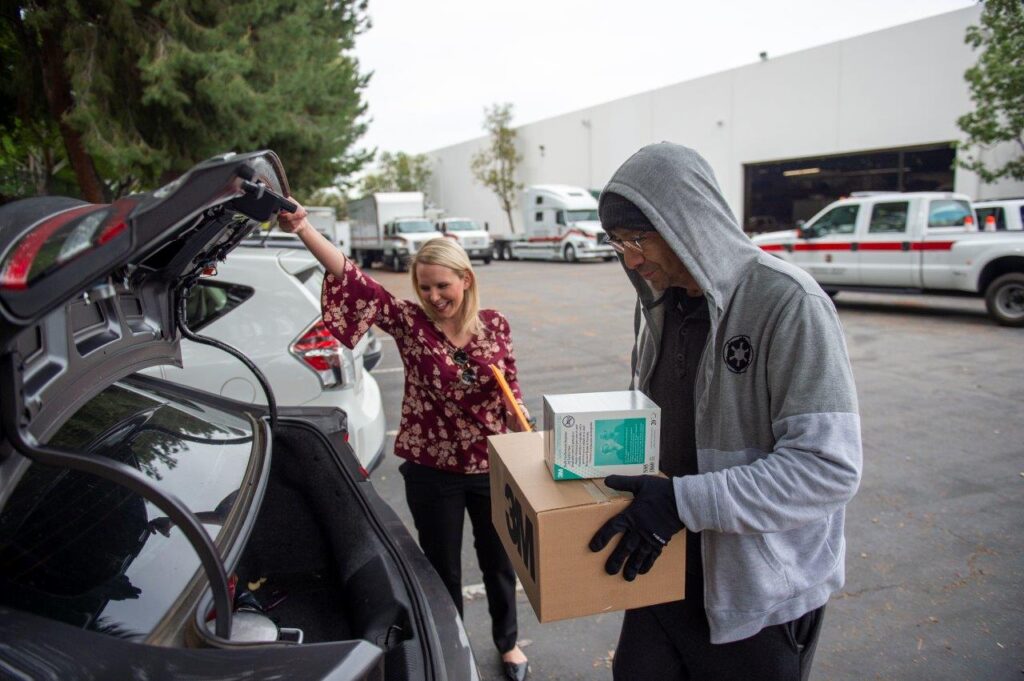 A woman assists a man loading a box of supplies into a vehicle, highlighting community support efforts during the pandemic.