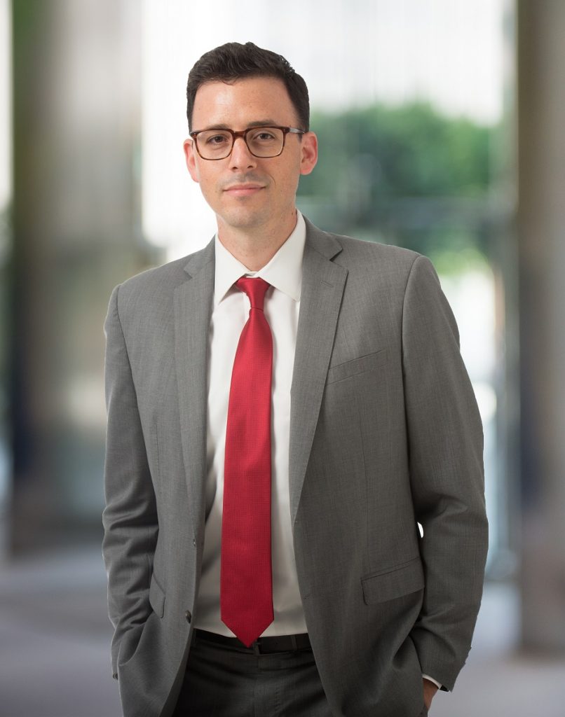 Professional portrait of a man in a gray suit and red tie, standing confidently in an office setting.