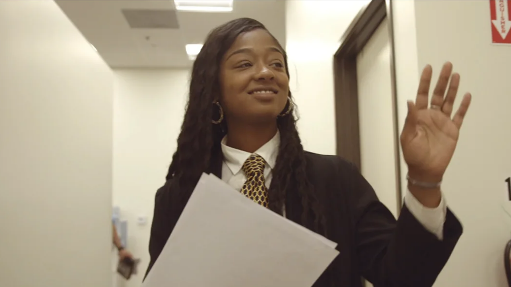A professional woman in a suit holding papers, smiling and waving while walking through a hallway.