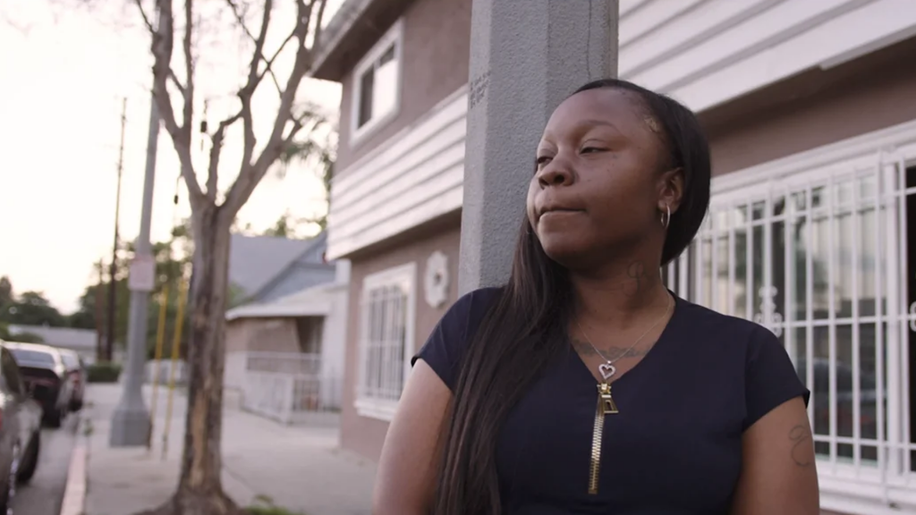 A woman standing by a pole outside a residential building, looking thoughtfully into the distance.