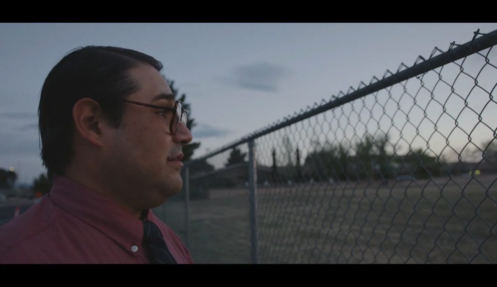 A man in glasses stands by a chain-link fence, looking contemplatively towards a field at dusk.