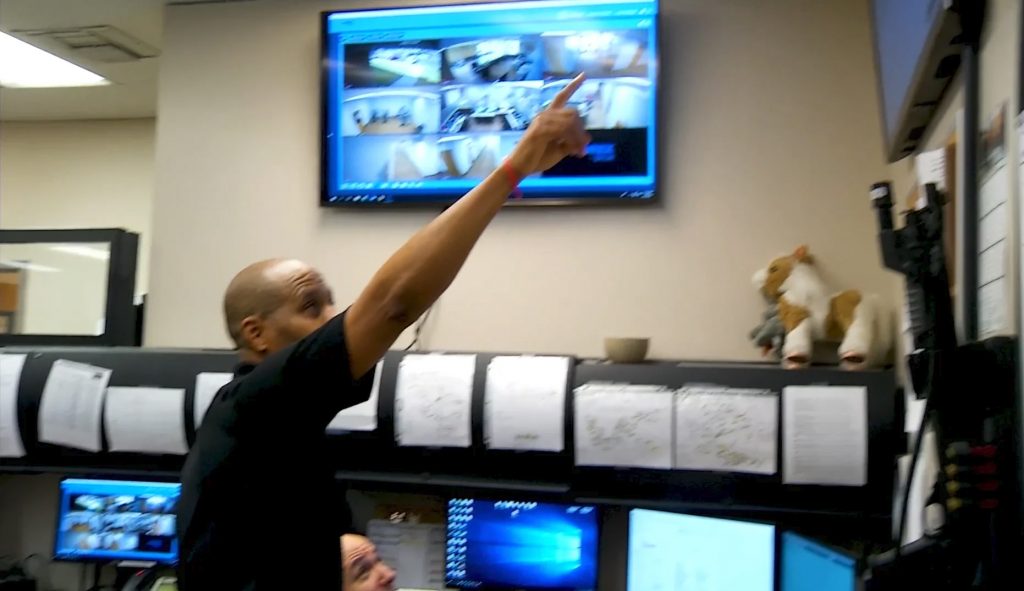 A staff member points at a monitor displaying multiple security camera feeds in a COVID-19 response command center.